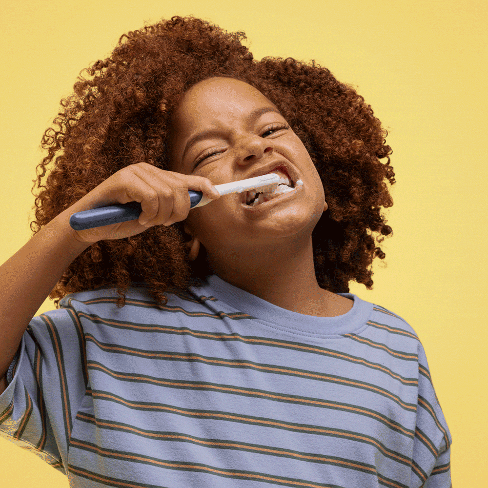 A child brushing their teeth with a blue quip Kids Sonic toothbrush, smiling and scrubbing enthusiastically against a yellow background.