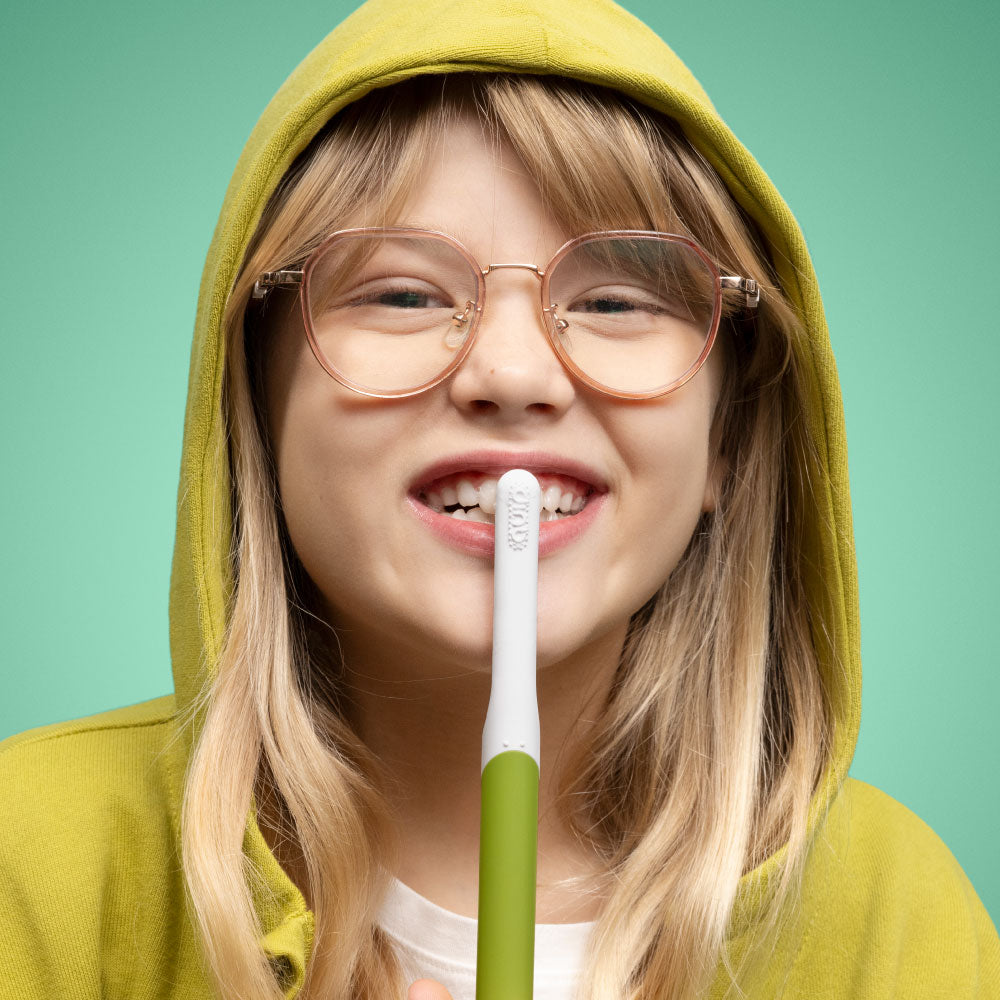 Person wearing a yellow hoodie and glasses, holding a green and white toothbrush against a green background