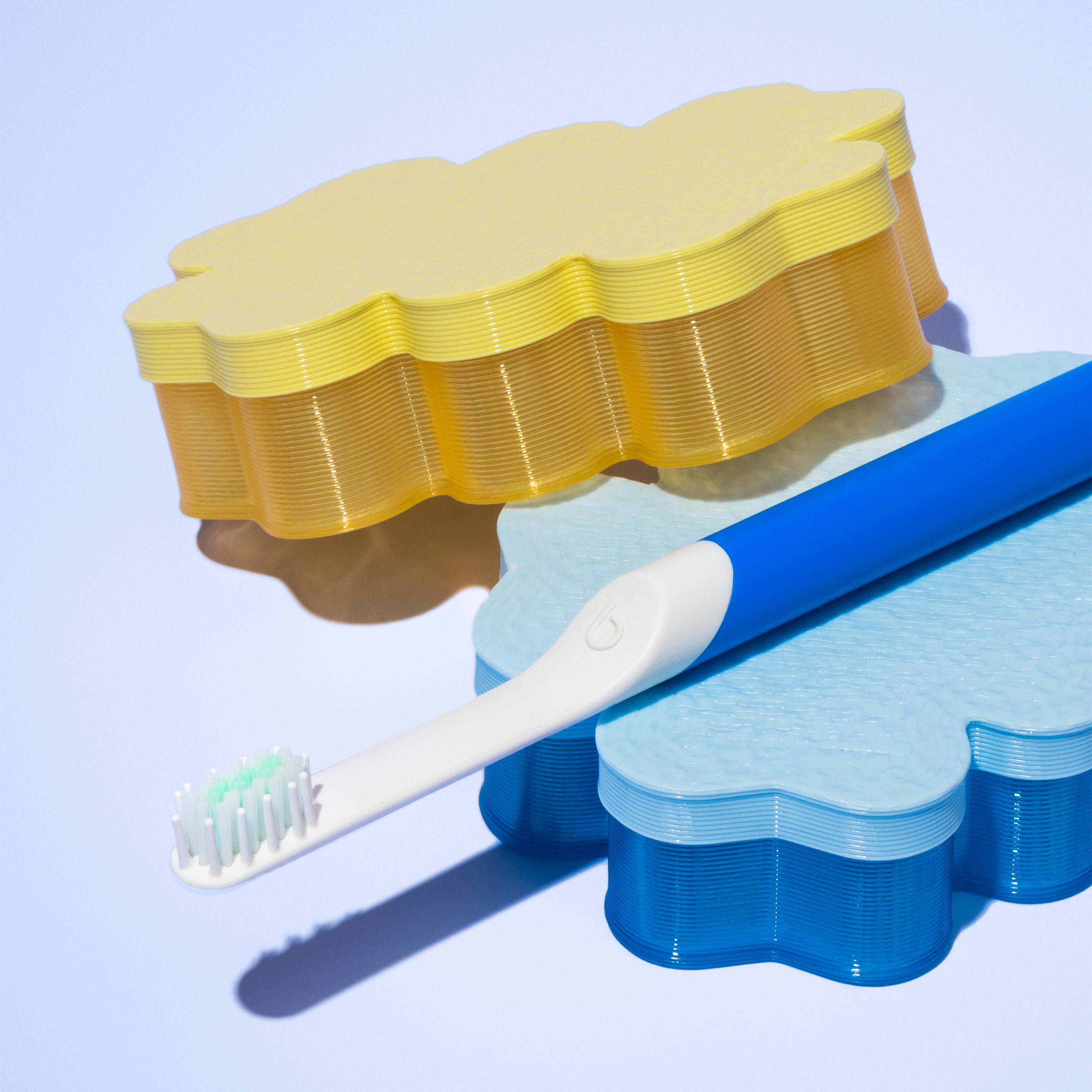 A blue quip Kids Sonic toothbrush resting on stacked cloud-shaped trays in yellow and blue, set against a soft light blue background.