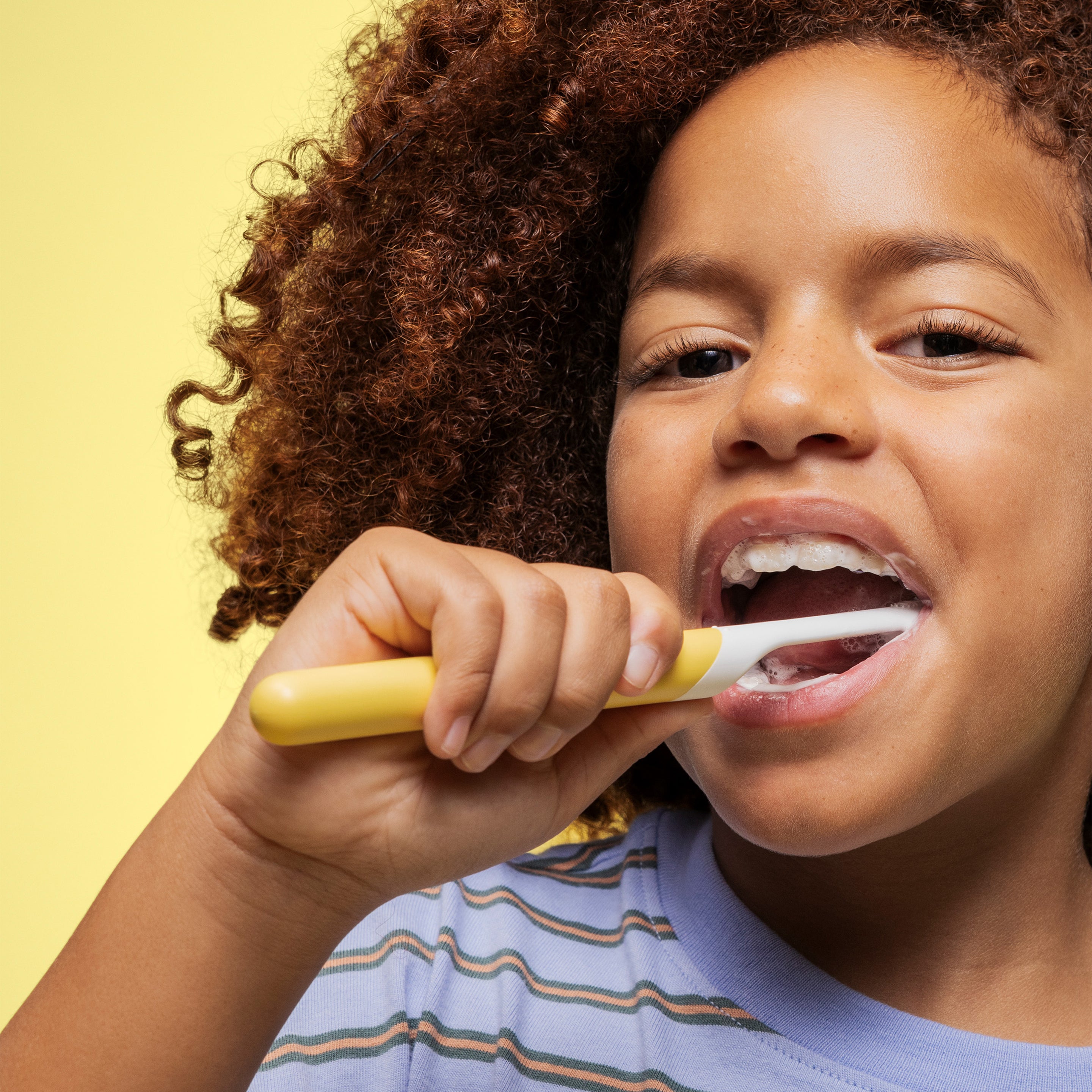 A child brushing their teeth with a yellow quip Kids Sonic toothbrush, smiling slightly while wearing a striped shirt against a yellow background.