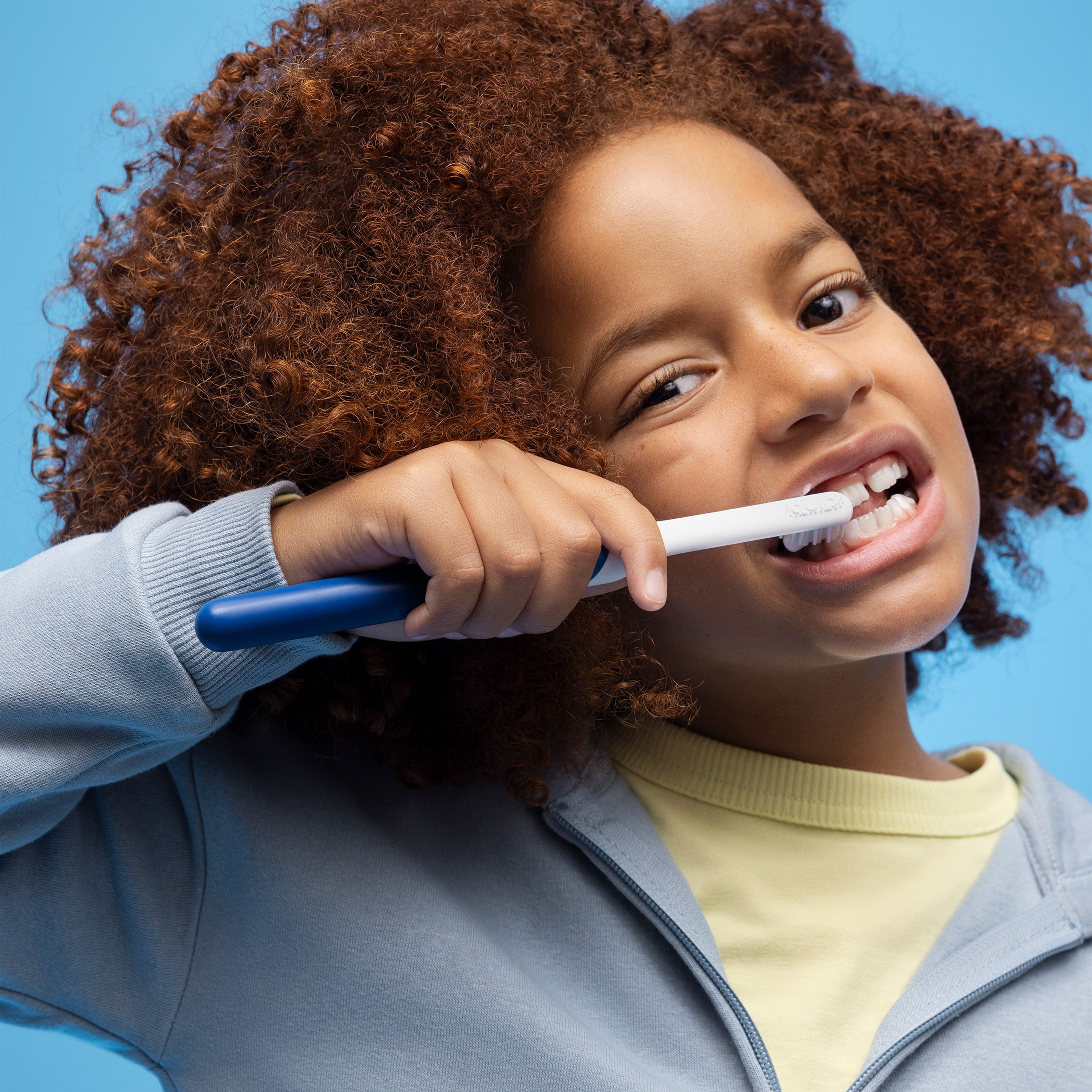 A child with curly hair brushing their teeth using a blue and white quip Kids Sonic toothbrush, smiling slightly against a bright blue background.