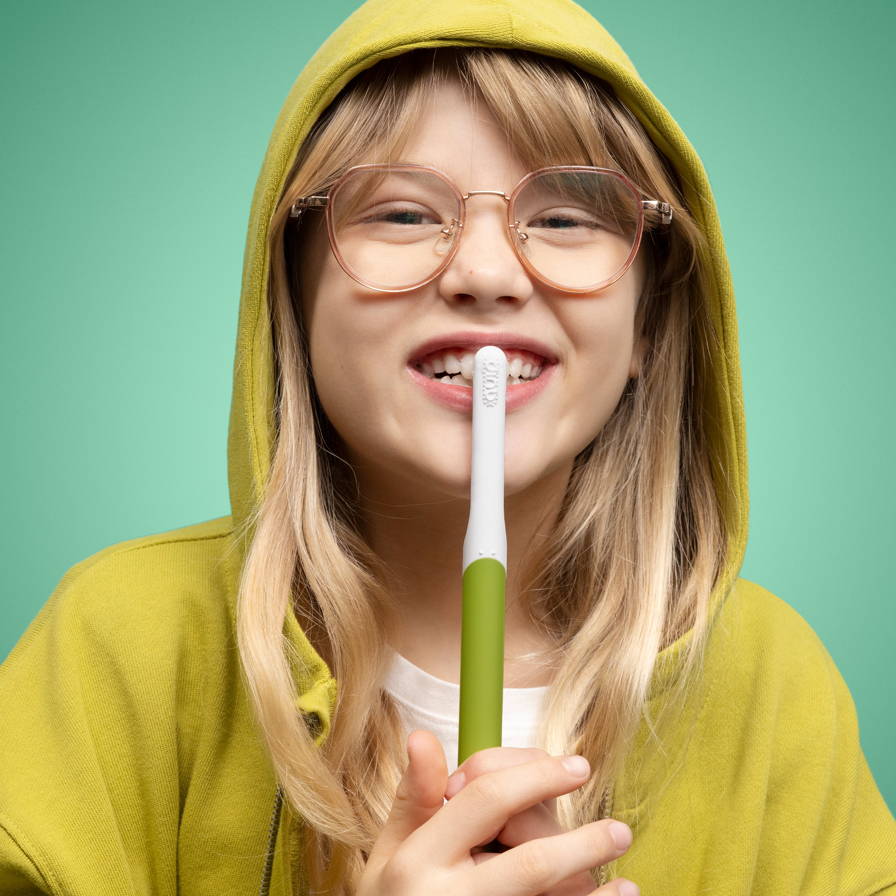 A child wearing glasses and a green hoodie smiles while holding a green and white quip Kids Sonic toothbrush upright in front of their mouth against a green background.