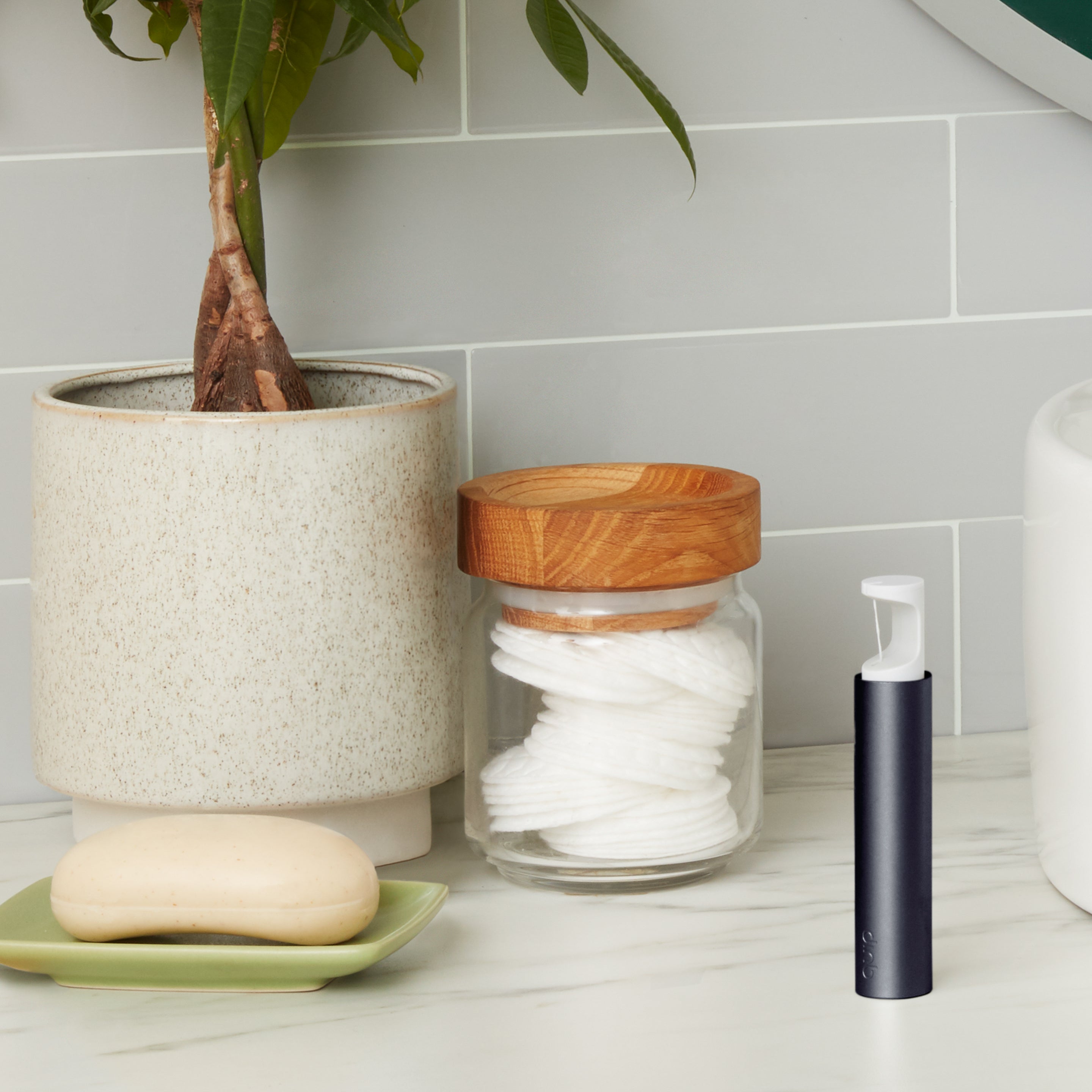 A sleek navy sliding floss dispenser standing upright on a bathroom countertop next to a potted plant, a jar of cotton pads with a wooden lid, and a soap dish.