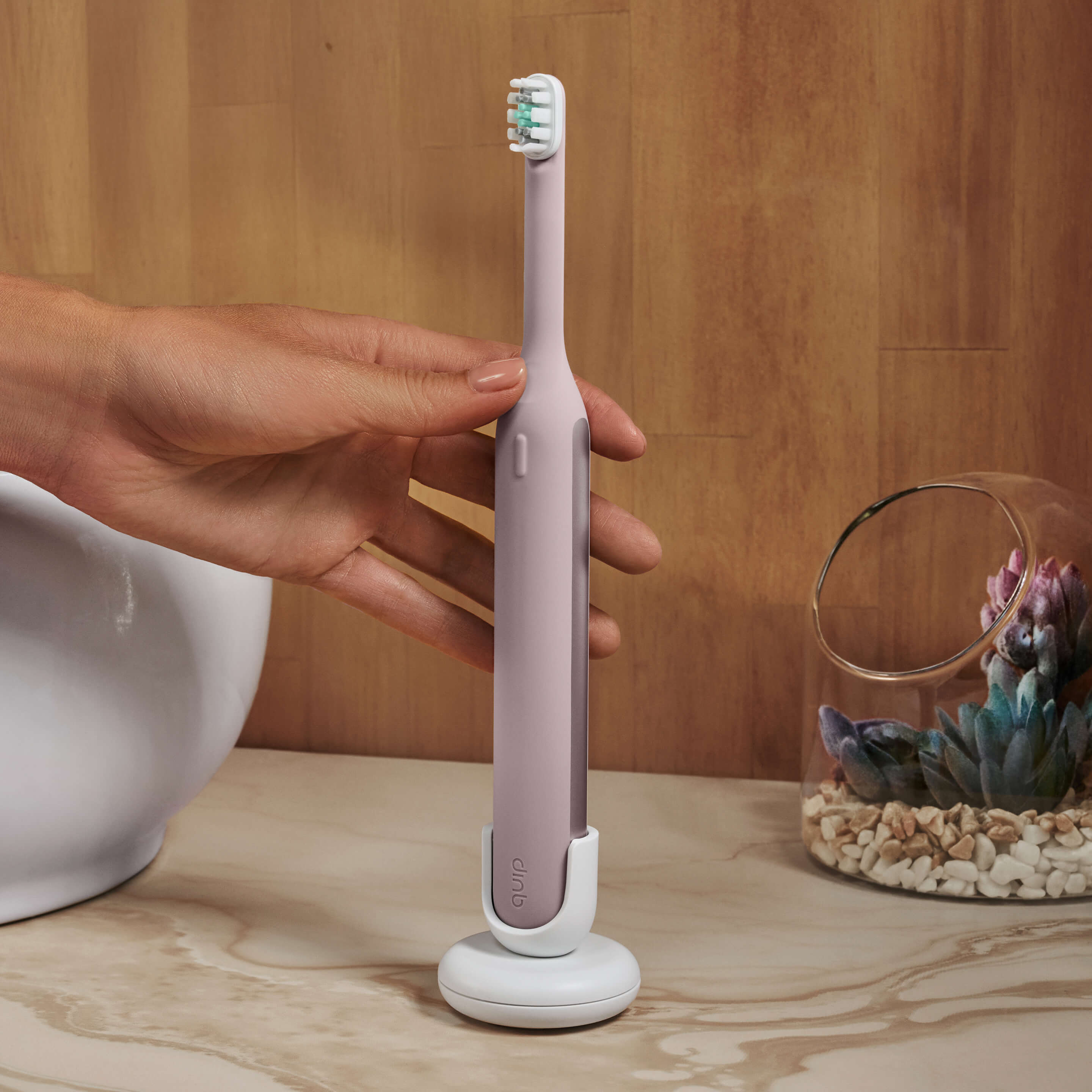 Hand placing a pink electric toothbrush onto its white charging stand on a bathroom counter.