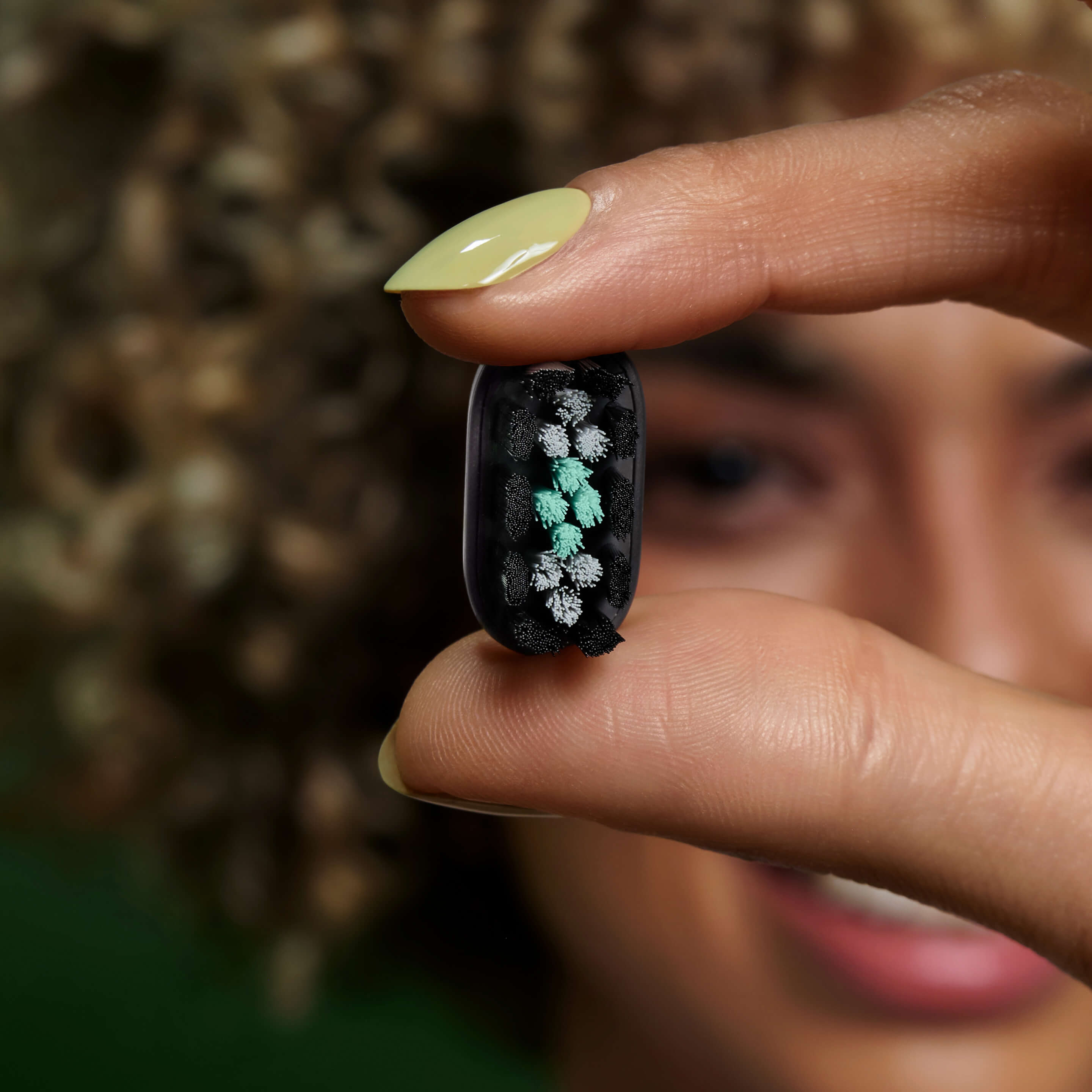 Person holding a small oval brush head between their fingers, showing its black and green bristles up close.