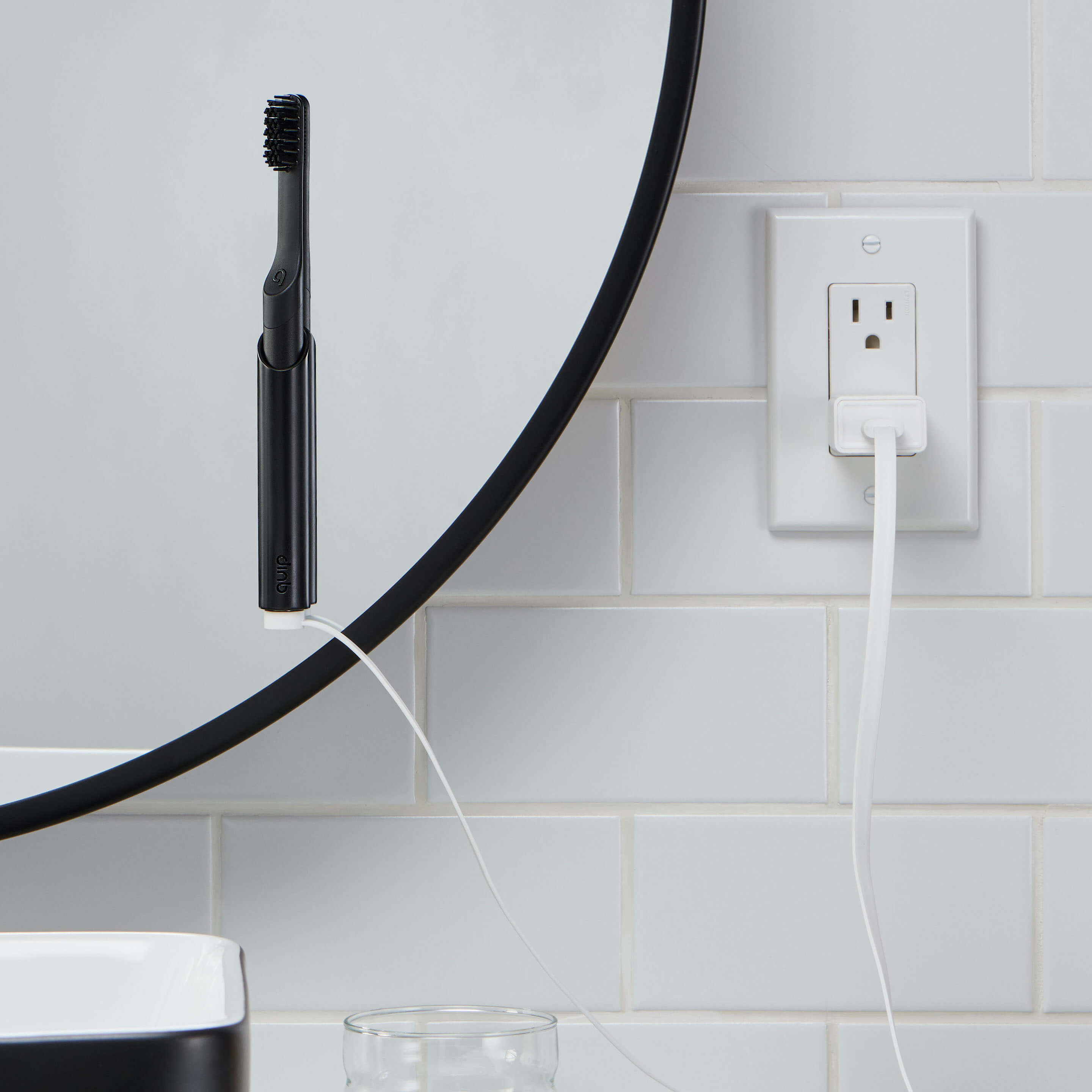 Black electric toothbrush with charging cable against a white tiled wall.
