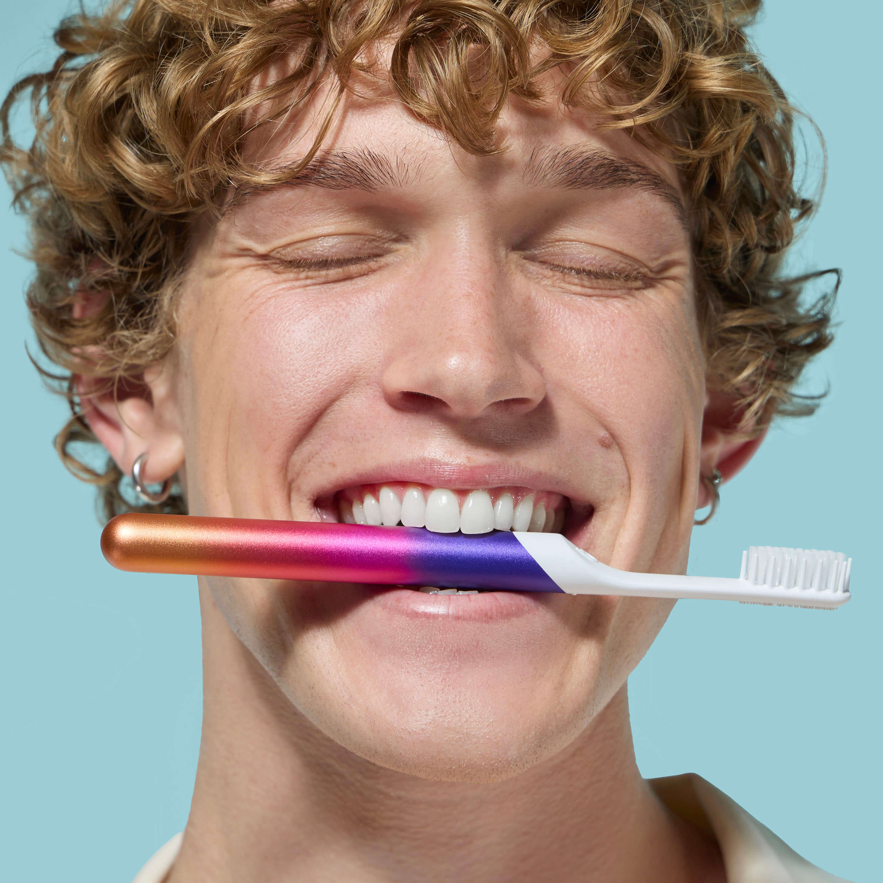 Smiling person with closed eyes holding a colorful gradient toothbrush between their teeth against a light blue background.