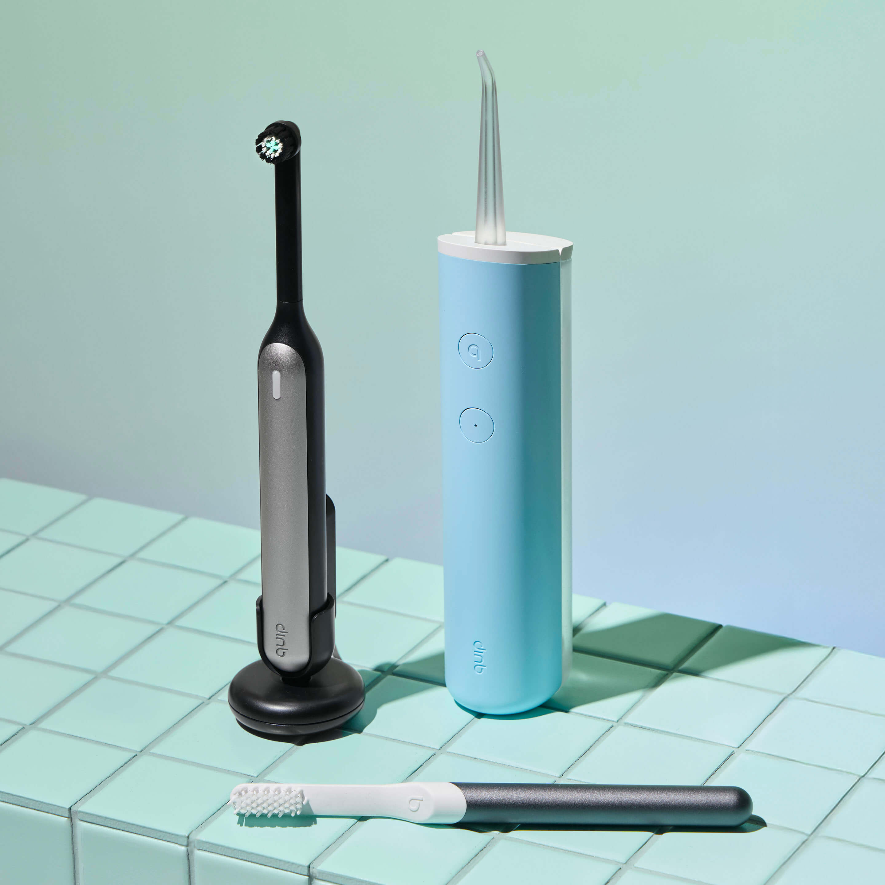 Set of dental care devices including a charcoal-colored electric toothbrush on a stand, a light blue water flosser, and a gray-and-white manual toothbrush laid on a mint-green tiled surface against a soft gradient background.