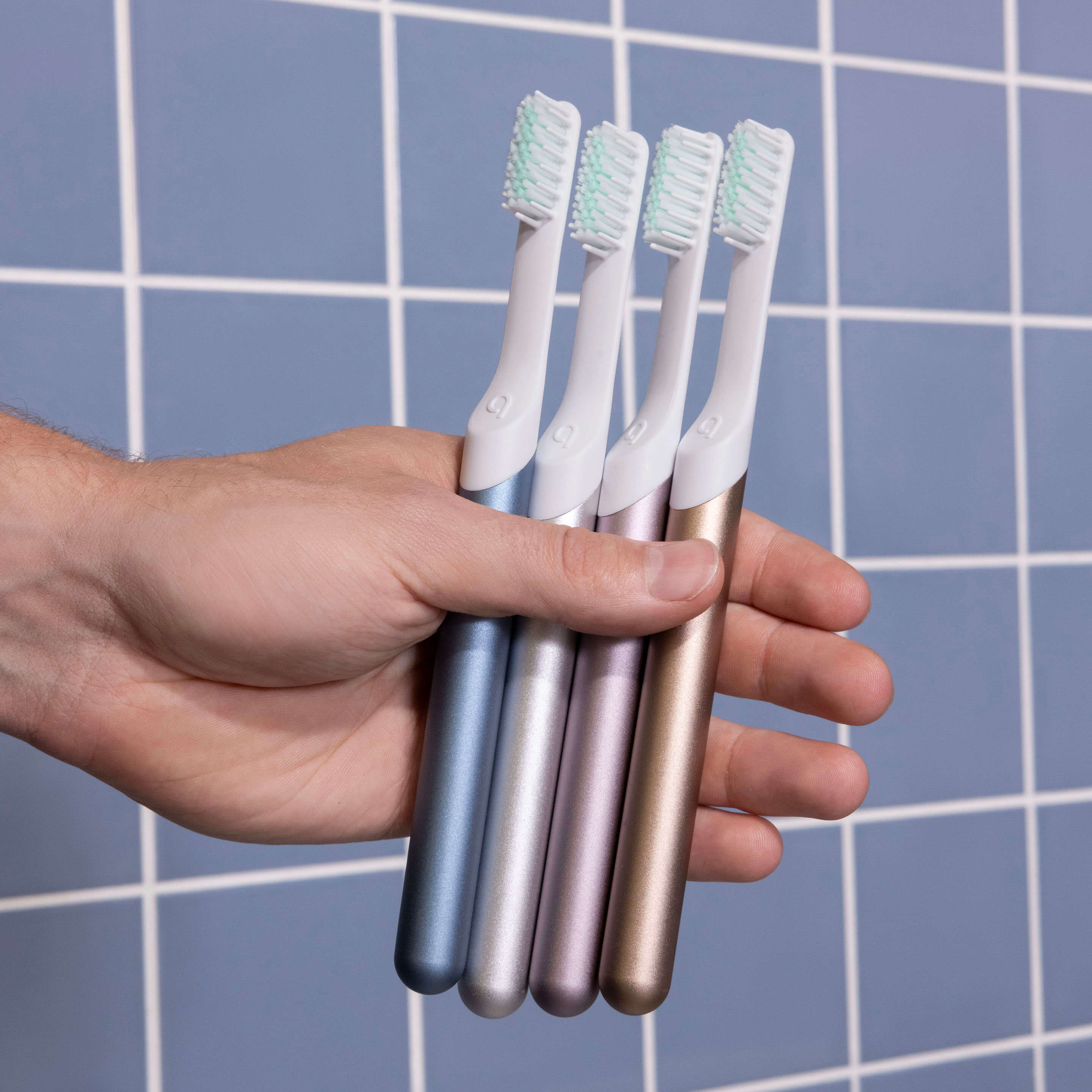 Hand holding four metallic-finish toothbrushes in blue, silver, rose, and gold colors, with white brush heads, in front of a blue tiled wall.