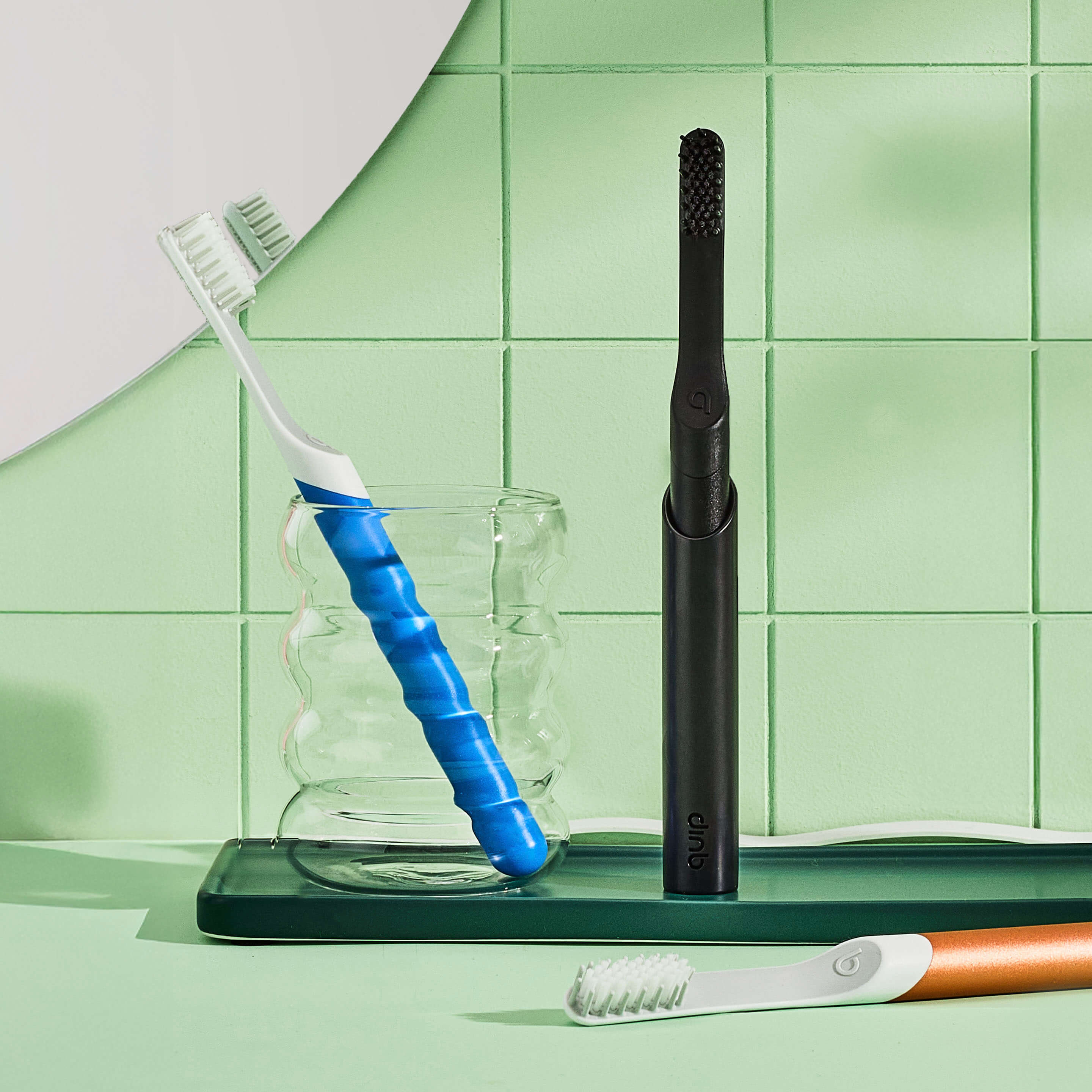 Sonic Toothbrush inside a glass with Black sonic toothbrush standing on the side and a green toothbrush laying down in a bathroom background with mirror.