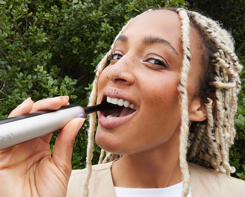 Smiling woman brushing her teeth outdoors with a silver quip electric toothbrush.