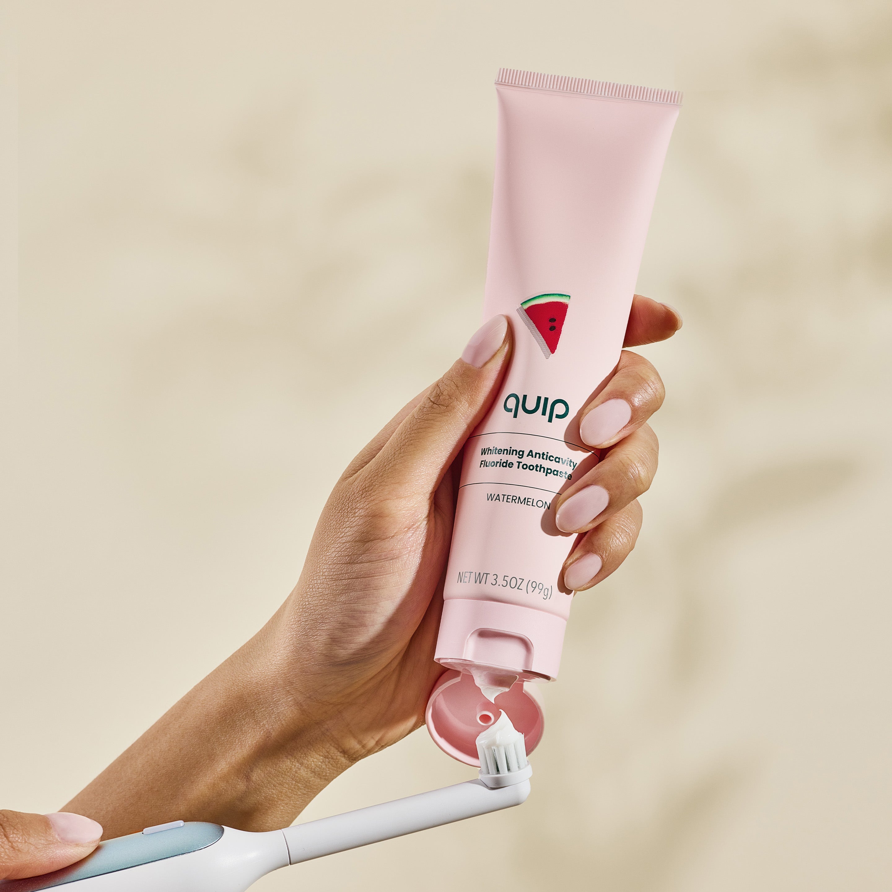 Hand holding a pink 'quip' toothpaste tube with a watermelon flavor, against a beige background.