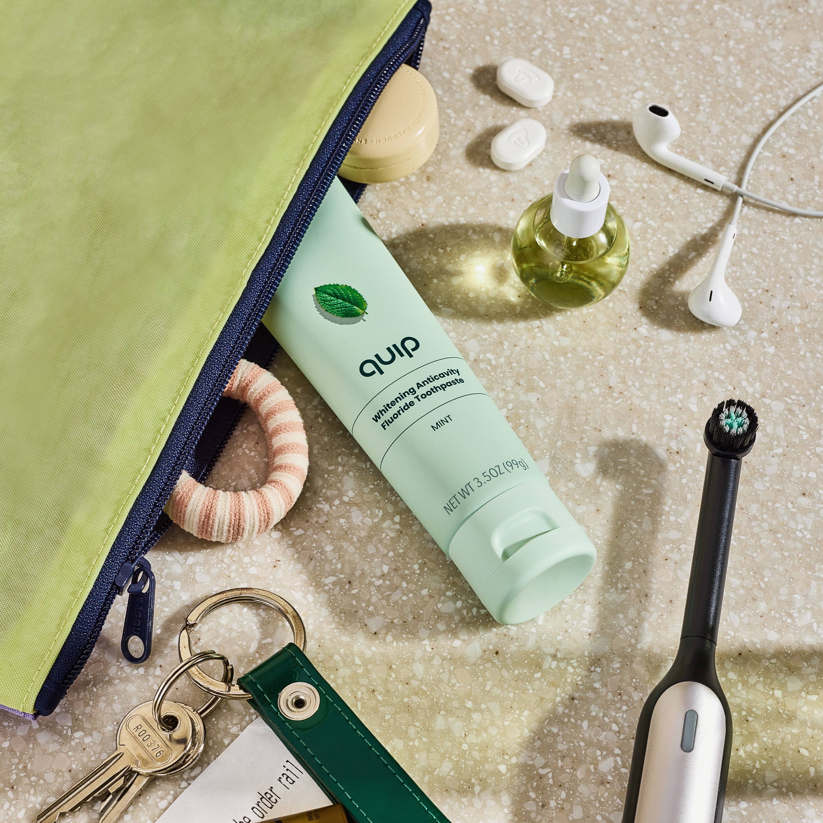 Flat-lay of a mint quip toothpaste tube with travel items including a zipper pouch, hair tie, keys, earbuds, and an electric toothbrush.
