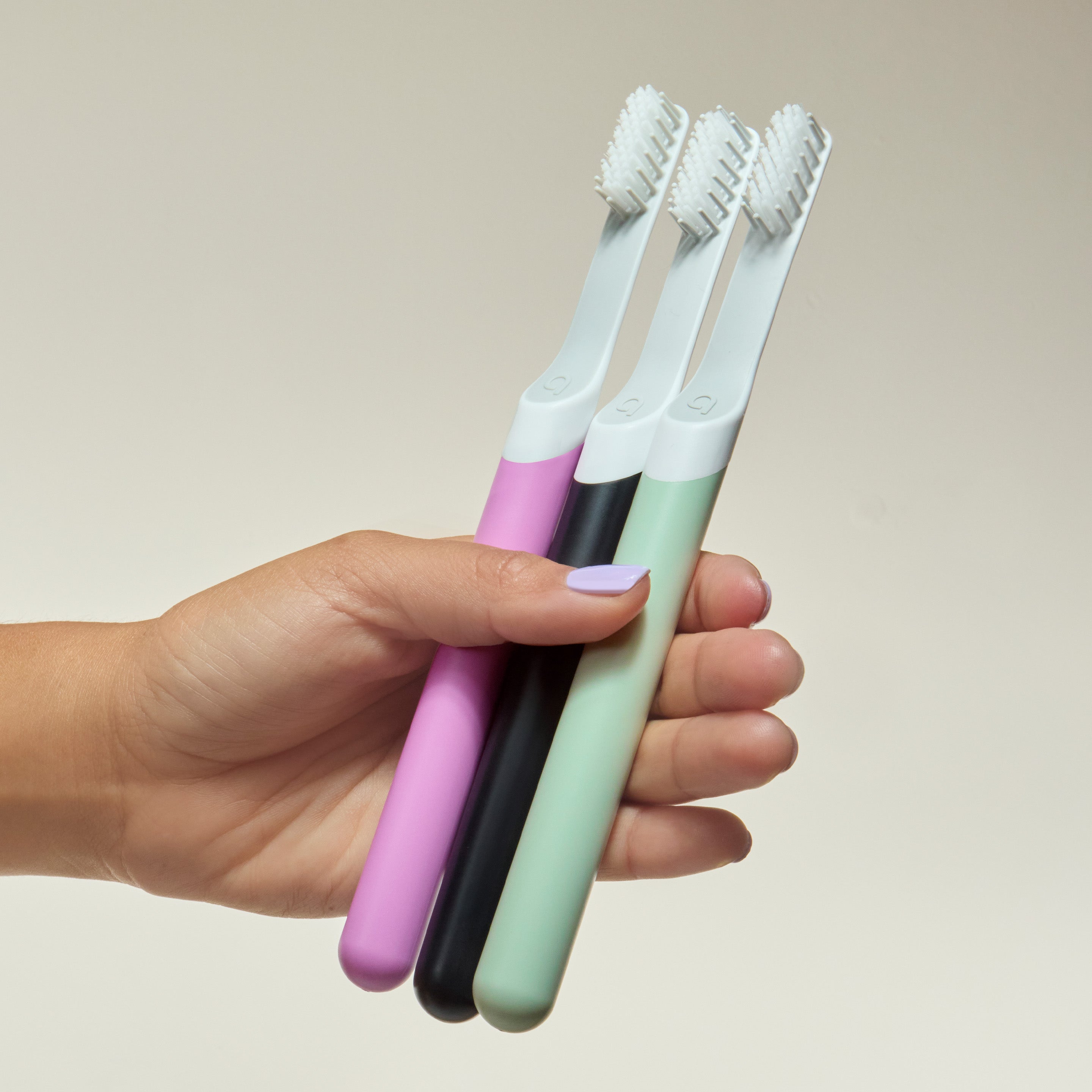 Hand holding three soft-bristle toothbrushes in pastel pink, black, and mint green.