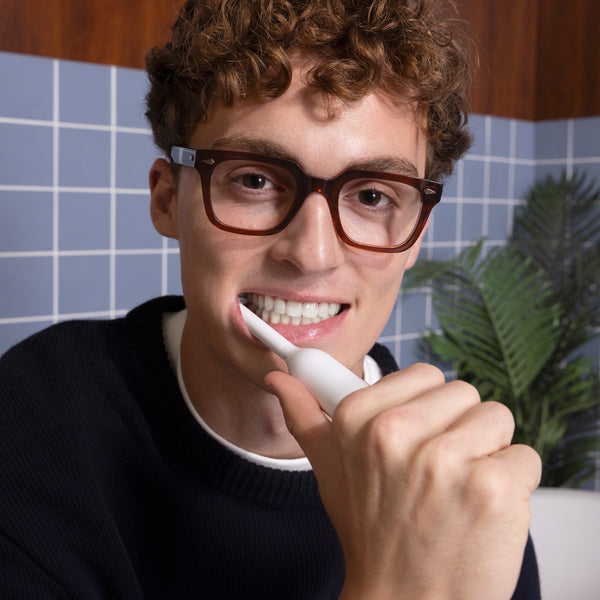 A man with curly brown hair and glasses brushing his teeth with a white quip electric toothbrush, with a blue tiled bathroom wall in the background.