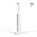 A white quip Rev Electric Toothbrush in the color Silver Cloud on its magnetic charging base, attached to a wall mount, with an ADA Accepted logo in the top right corner.