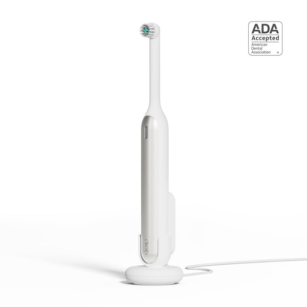 A white quip Rev Electric Toothbrush in the color Silver Cloud on its magnetic charging base, attached to a wall mount, with an ADA Accepted logo in the top right corner.