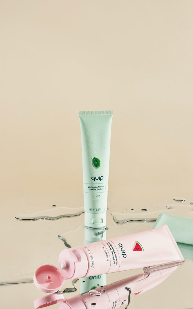 Quip toothpaste tubes in mint and watermelon flavors on a reflective wet surface with water droplets.