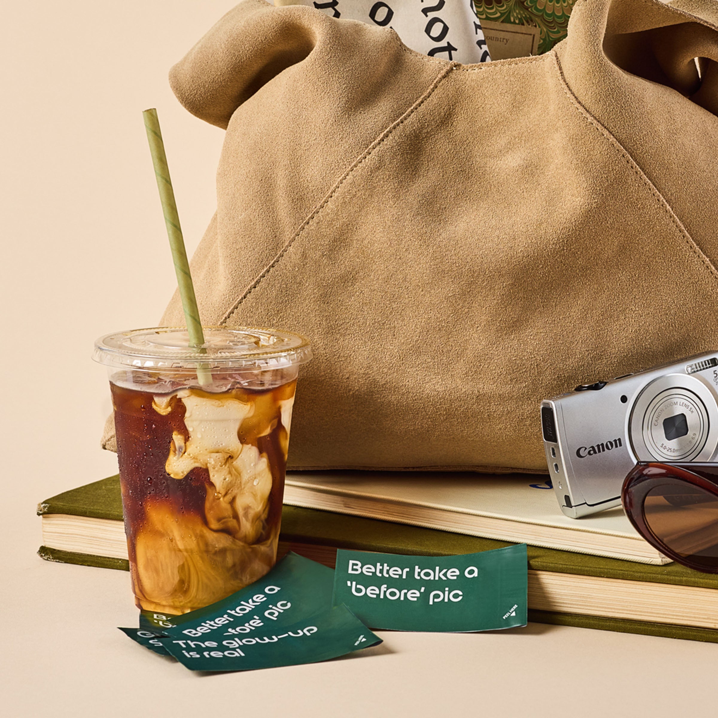 Beige handbag with a cup of iced coffee, camera, and sunglasses on books against a beige background