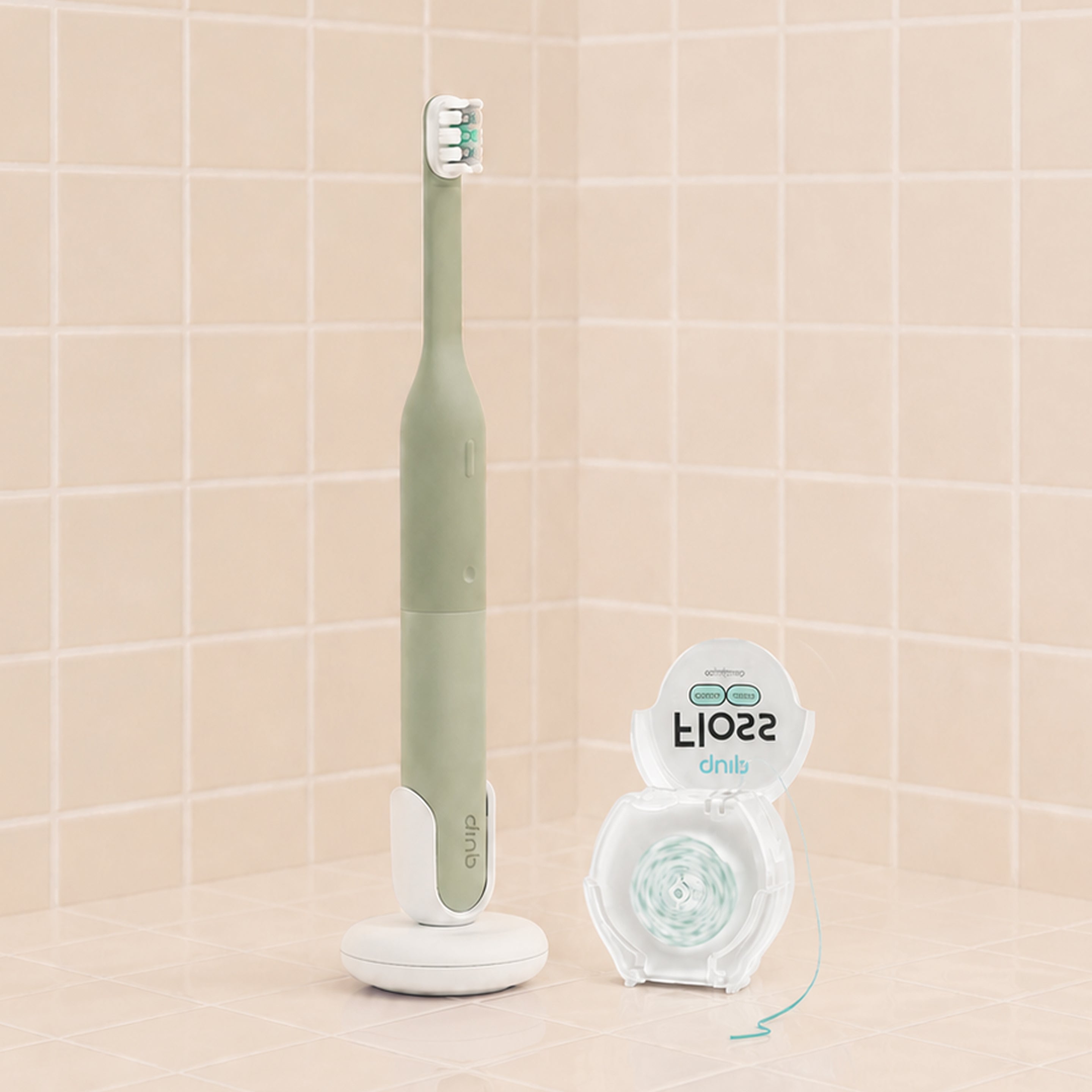 A mint green and white electric toothbrush standing upright on a charging base next to a clear container of dental floss, set against a beige tiled background.