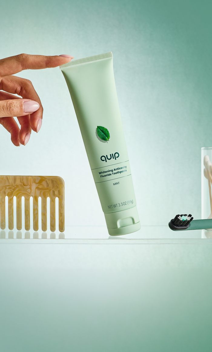 A hand holds a tube of Quip Whitening Baking Soda Toothpaste against a mint green background, with a wooden comb and toothbrush visible on a shelf below.